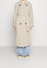 Beige trenchcoat van een wolmengsel met een dubbelknopenontwerp, zwarte knopen, grote zakken en een bindceintuur, gecombineerd met lichtblauwe lose spijkerbroek.