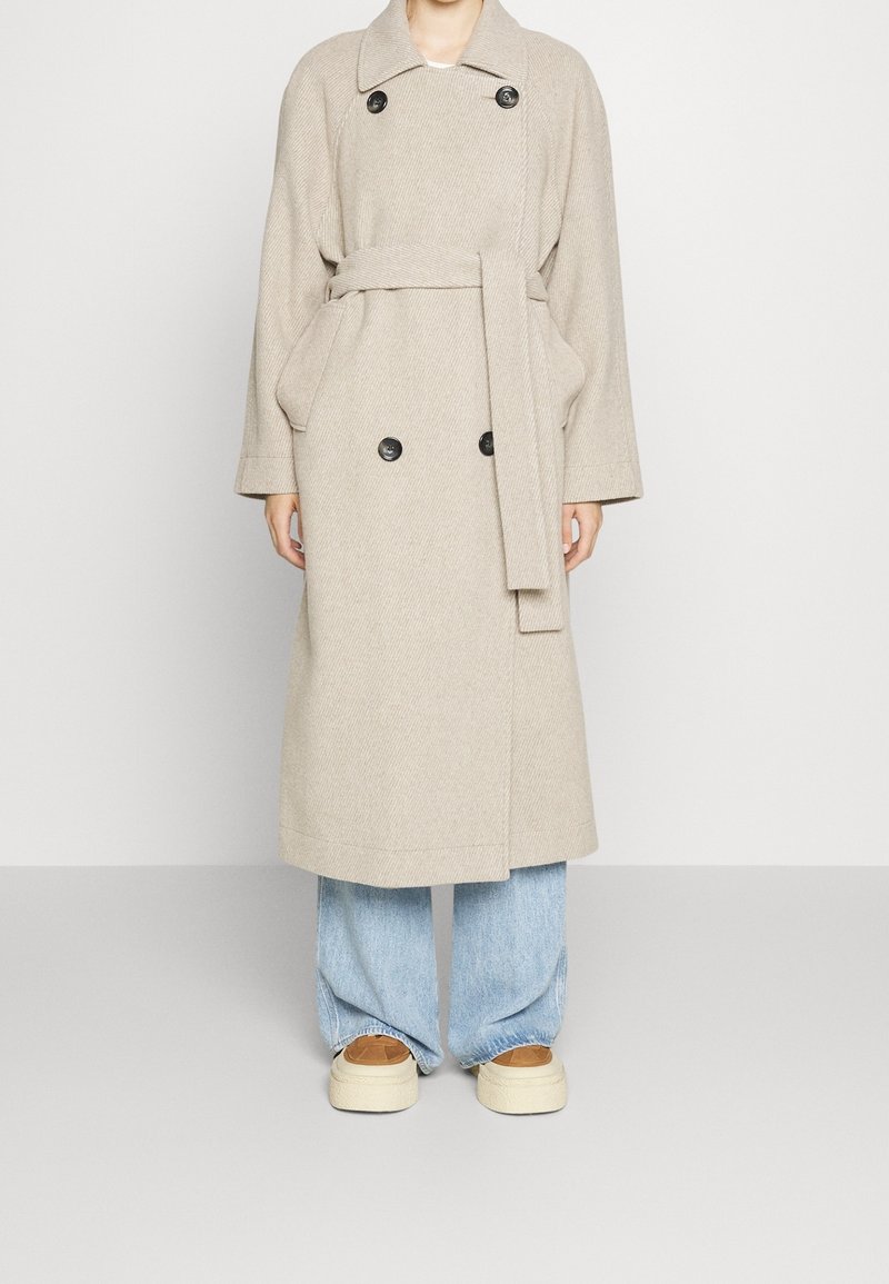 Beige trenchcoat van een wolmengsel met een dubbelknopenontwerp, zwarte knopen, grote zakken en een bindceintuur, gecombineerd met lichtblauwe lose spijkerbroek.