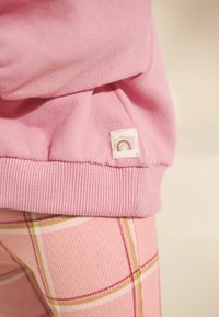 Tissu de sweat-shirt rose avec un ourlet côtelé, doté d'une petite étiquette avec un design de rainbow. Associé à un legging à motif tartan rose.