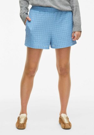 Shorts tissés bleu clair avec une taille élastique et des poches latérales. Le tissu a un motif texturé, complété par des baskets beiges.