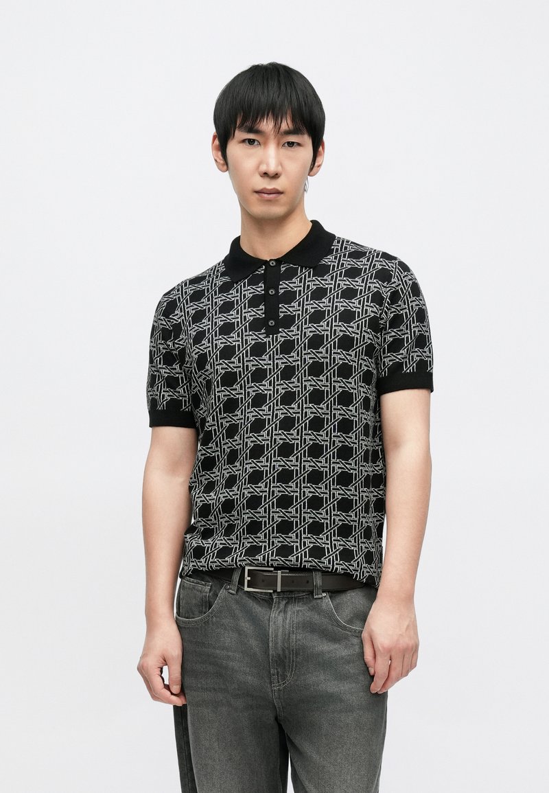 Čierne polo tričko s kontrastným geometrickým bielym vzorom, krátkymi rukávmi a pevným čiernym golierom, nosené s tmavosivými džínsami z denimu.