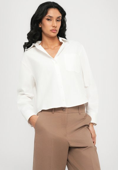 Chemise blanche courte à boutons avec une poche poitrine unique, associée à un pantalon beige taille haute, présentant une coupe droite et une texture lisse.