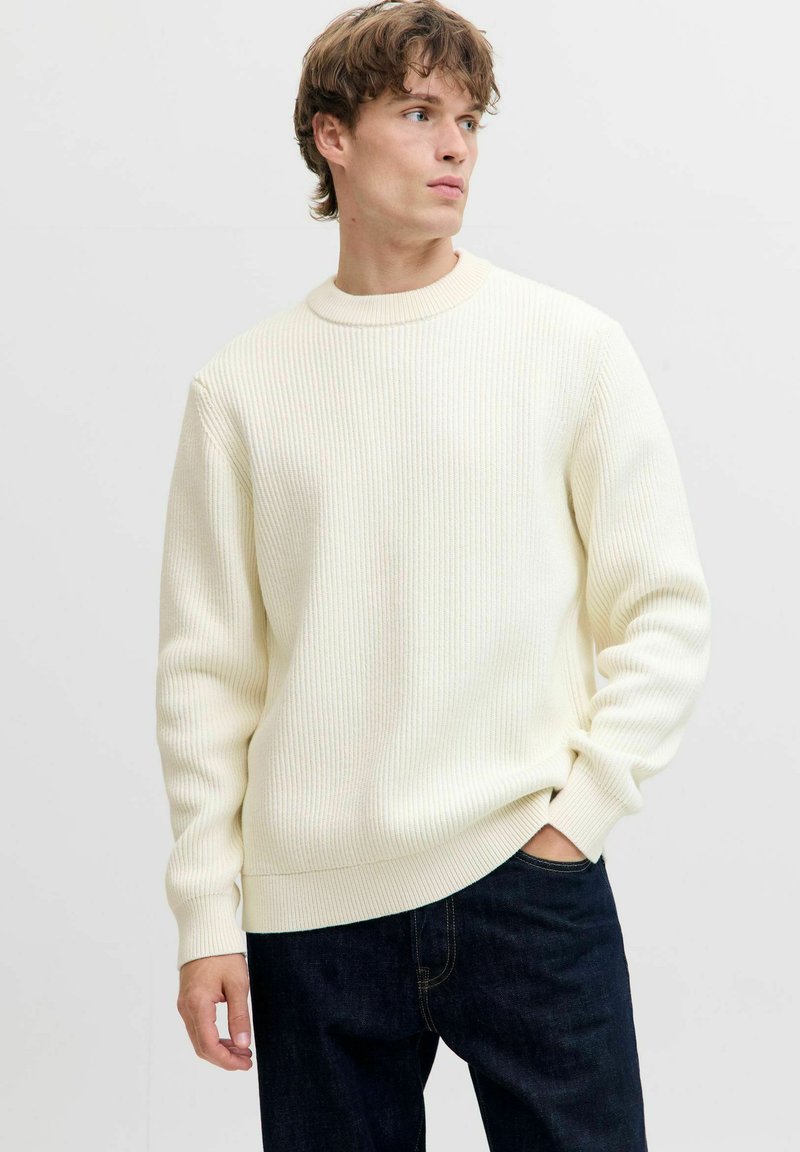 Maglione a coste color crema con scollatura a girocollo e maniche lunghe, abbinato a jeans scuri. La texture sembra morbida e leggermente elasticizzata.