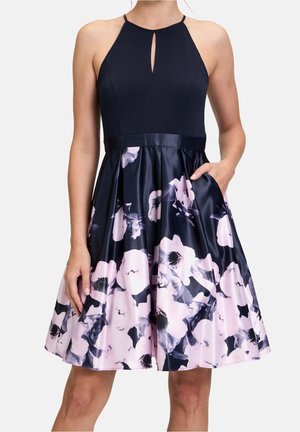 Robe noire sans manches avec encolure fendue et jupe en satin ornée d'un grand imprimé floral rose et noir, modèle avec une main dans la poche.