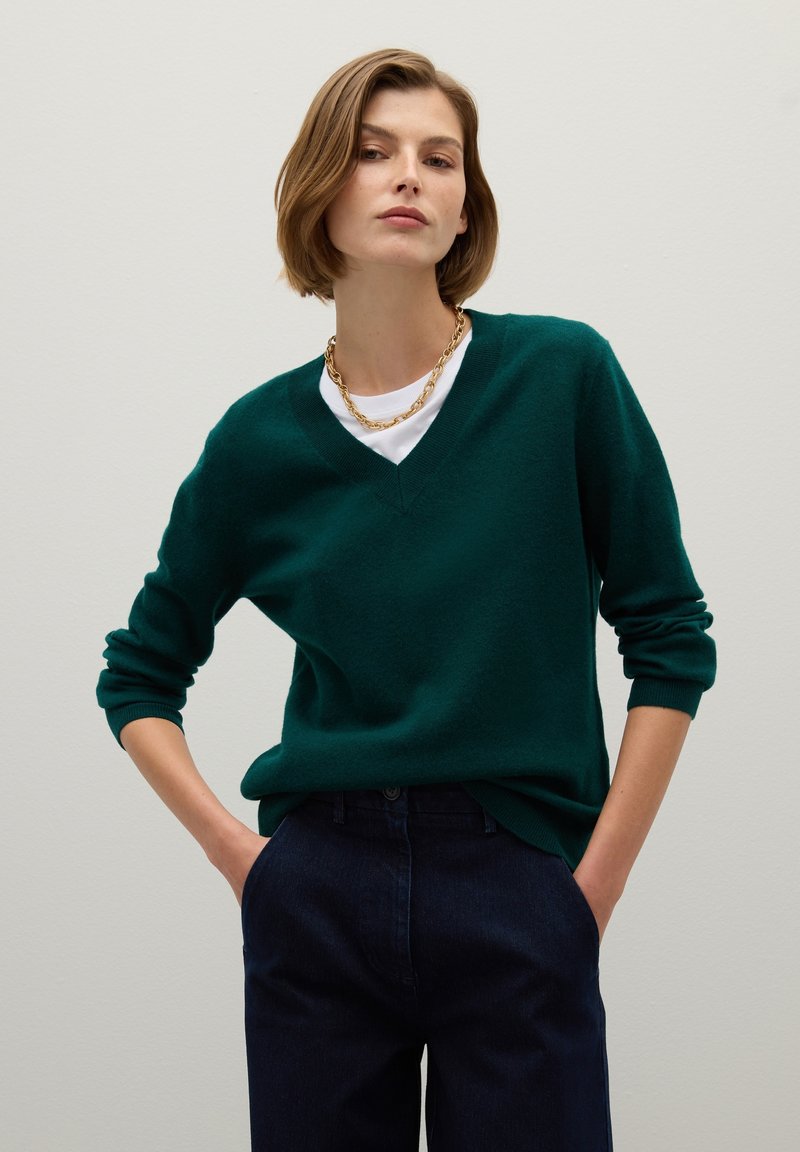 Suéter verde de cuello en V hecho de una tela suave y texturada, con mangas largas y un corte relajado, combinado con pantalones de mezclilla oscuros.