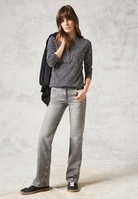 Haut à manches longues noir avec un motif de feuilles blanches, associé à un jean évasé gris clair et des baskets noires. La silhouette se tient devant un fond neutre.