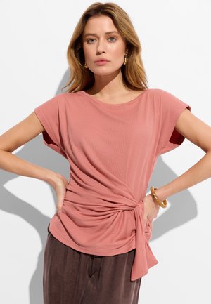 Kobieta o jasnobrązowych włosach, ubrana w różową bluzkę z krótkim rękawem i wiązaniem po boku oraz ciemnobrązowe spodnie, stoi z rękami na biodrach na białym tle.