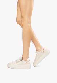 Zapatillas de deporte color beige claro con una textura de cuero suave, que presentan adornos decorativos en dorado y una suela de plataforma blanca elevada.