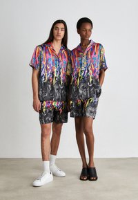 Camicie e pantaloni corti abbinati da uomo e da donna con motivi a vortice multicolori su sfondo nero, realizzati in tessuto leggero.