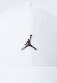 Hvid baseballkasket lavet af tekstureret stof, med et sort Jumpman-logo på fronten. Bøjelig skygge og syede detaljer er til stede.