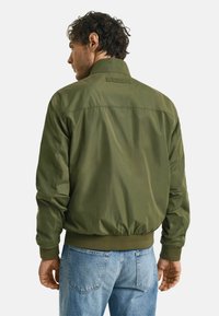Giacca bomber verde oliva con colletto alto, polsini e orlo elasticizzati. Presenta una texture liscia e un design pulito senza hardware visibile.