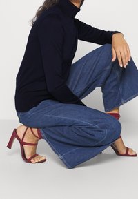 Laivastonsininen poolopaita, istuva malli; leveälahkeiset tummat farkut näkyvillä saumoilla; viininpunaiset remmikkäät korkokengät.