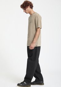 Kortærmet, beige bomulds skjorte med et lille logo; stylet med sorte, løstsiddende bukser og sorte sko. Rent, enkelt design.