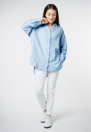 Lichtblauwe oversized button-up shirt in textielmateriaal, gecombineerd met witte slim-fit broek en witte sneakers met blauwe accenten.