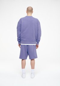 Uomo con testa rasata che indossa un felpa viola e pantaloni corti abbinati, calze bianche e scarpe da tennis, sta in piedi di spalle su uno sfondo bianco.