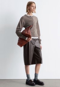 Brauner gestrickter Cardigan mit rundem Ausschnitt und Knopfverschluss, kombiniert mit einem braunen Leder-Midirock. Hält eine strukturiert rötlich-braune Handtasche.