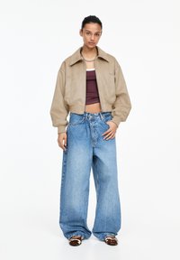 Beige kort jacka, vinröd linne och ljusblå vida jeans med en blekt finish. Brun och vit fotbeklädnad ger kontrast.