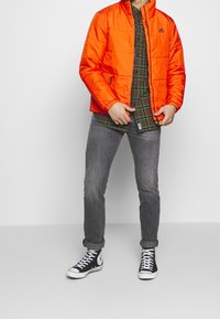 Chaqueta acolchada naranja brillante con un cuello alto, combinada con una camisa de cuadros verde y jeans grises. Zapatillas altas negras como calzado destacado.