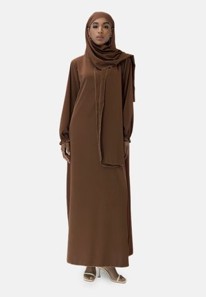 Vestito lungo marrone con maniche, abbinato a un hijab, realizzato in tessuto fluido. Presenta una leggera lucentezza e dettagli dorati sui bordi. Indossato con tacchi di colore chiaro.
