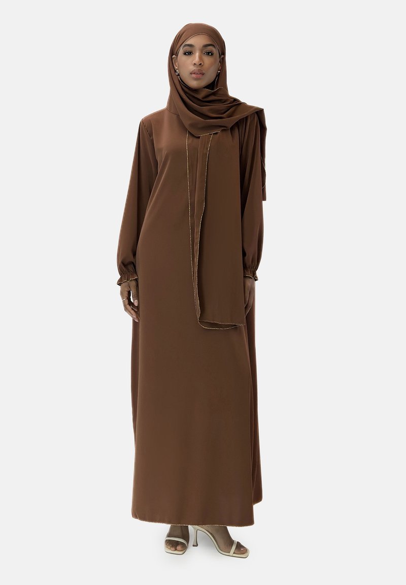 Brun langærmet kjole med en matchende hijab, lavet af flydende stof. Har en subtil glans og guldkanter. Båret med lyse hæle.
