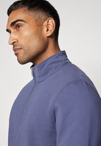 Muž s krátkymi tmavými vlasmi a strniskom, ktorý má na sebe modrý pulóver so zipsom na štvrť, sa pozerá doľava na pozadí v svetlej farbe.