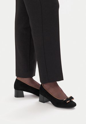 Schwarze Wildleder-Pumps mit kleinem schwarzem Schleifchen und beigem Rand, getragen mit dunklen feinen Strumpfhosen und eng anliegenden schwarzen Hosen.