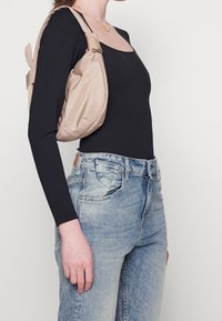 Bolso bandolera beige con un acabado suave y texturizado, llevado sobre el hombro. Blusa negra de manga larga combinada con jeans de mezclilla azul claro.