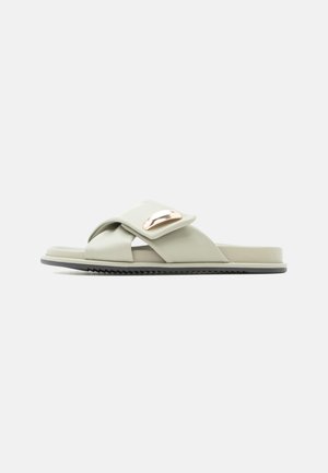 Sandalo slip-on verde chiaro con ampie fasce incrociate e un accento ovale metallico sulla parte superiore, suola piatta con fondo strutturato.