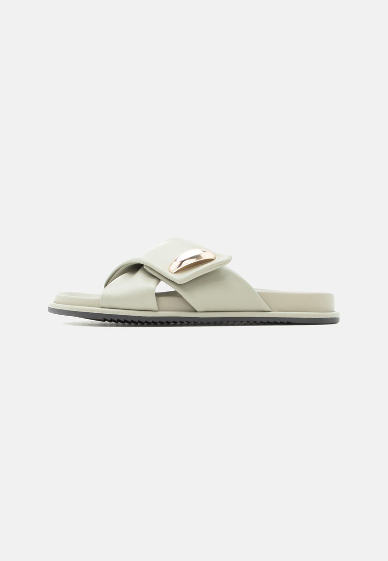 Ανοιχτό πράσινο σανδάλι τύπου slip-on με φαρδιές λουριά που διασταυρώνονται και μεταλλικό οβάλ διακοσμητικό στο πάνω μέρος, με επίπεδη σόλα και υφή στο κάτω μέρος.