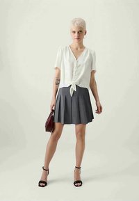 Weiße kurzärmlige Bluse mit einer durchgehenden Knopfleiste und gebundenem Saum, kombiniert mit einem grauen, hoch taillierten, gefalteten Rock und schwarzen, hochhackigen Sandalen.