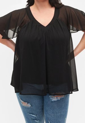 Frau trägt eine lockere schwarze transparente Bluse mit flatternden Ärmeln und blaue zerrissene Jeans, steht mit den Händen in der Hüfte vor weißem Hintergrund.