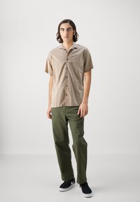 Camisa de cuadro de manga corta, en beige y marrón, combinada con pantalones verde oliva. El calzado presenta zapatillas oscuras con detalles en blanco.