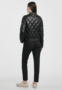 Czarna pikowana kurtka puchowa z wysokim kołnierzem, z diamentowym wzorem i ribbowanym dołem, sparowana z czarnymi spodniami i beżowymi trampkami.