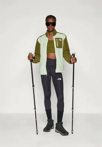 Kaki un zaļš flīsa jaka ar rāvējslēdzēja kabatu, saskaņota ar atbilstošu topi un tumšām legingām. Melni pārgājienu zābaki un trekinga nūjas.