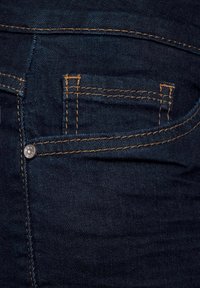 Tummansiniset farkut, joissa on takatasku ja oranssilla ompeleella korostetut yksityiskohdat sekä metallinen nappi. Kangas on sileä ja kestävä.