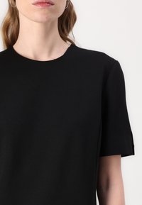 Crni kratki rukavi top s okruglim izrezom, glatkom teksturom i minimalističkim dizajnom. Suptilni detalj šivanja blizu izreza.