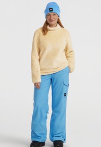 Fluffy lichtgele pullover, felblauwe broek met cargozakken, en een lichtblauwe gebreide beanie, gecombineerd met zwarte schoenen.