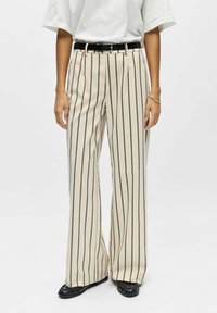 Pantalon beige à jambes larges avec des rayures verticales noires, doté d'une ceinture noire et d'une texture lisse. Porté avec des chaussures noires.