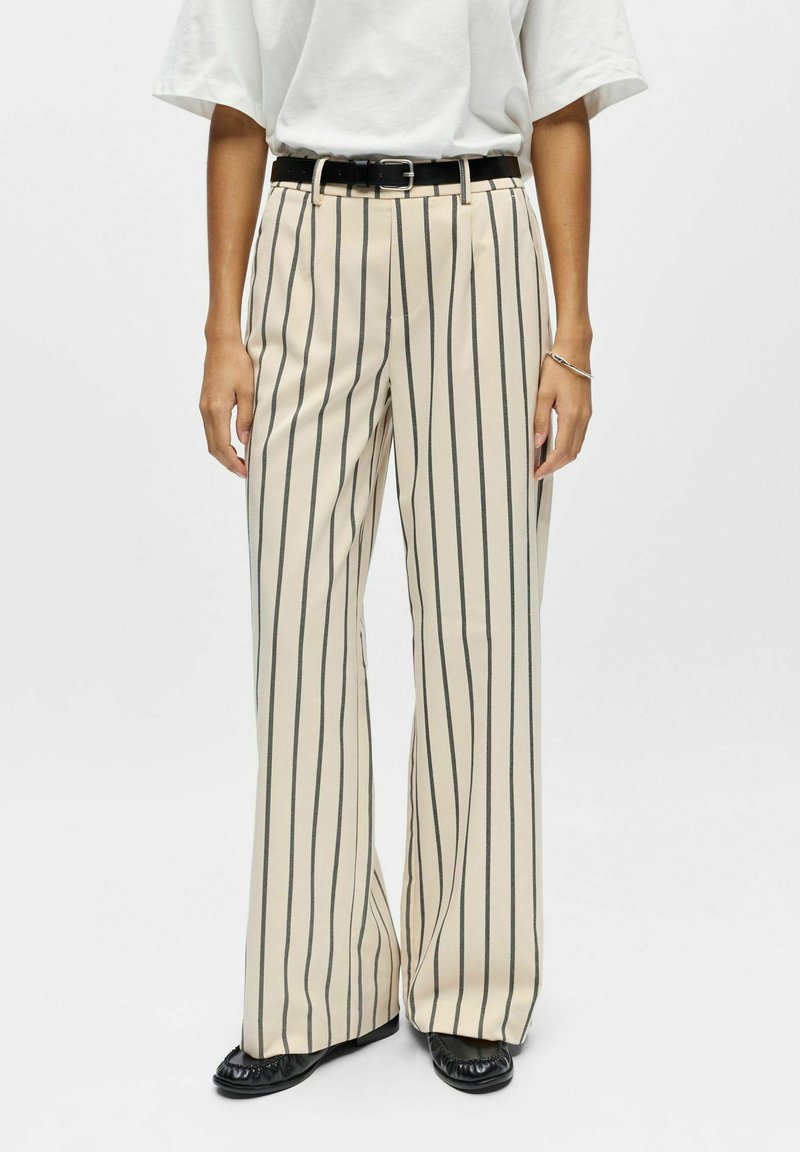 Pantalon beige à jambes larges avec des rayures verticales noires, doté d'une ceinture noire et d'une texture lisse. Porté avec des chaussures noires.