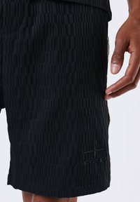 Black shorts featuring a textured, patterned fabric with vertical lines, side zipper pockets, and a stitched logo detail on the hem.
