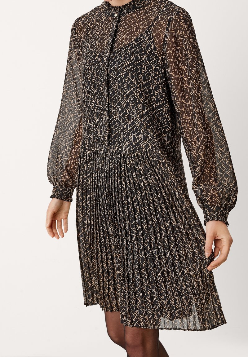 Robe noire semi-transparente à manches longues avec imprimé abstrait beige, jupe plissée et col haut avec fermeture par bouton à la gorge.
