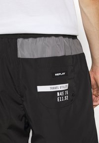 Shorts noirs avec des accents gris, taille élastique et une poche arrière avec une étiquette indiquant "TRAVEL UTILITY" avec des coordonnées.