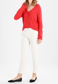 Czerwony sweter z dekoltem w kształcie litery V, długie rękawy; zestawiony z białymi dopasowanymi spodniami i czarnymi płaskimi butami. Prosty design i współczesna sylwetka.