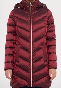 Burgundy puffer-takki, jossa on kevyet viivasto, vetskari edessä, huppu, sivutaskut ja kullanväriset yksityiskohdat. Valmistettu kiiltävästä, ”quilted” kankaasta.
