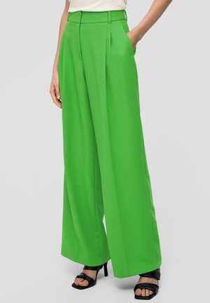 Pantalons larges verts vifs en tissu lisse, dotés de plis et de poches latérales. Portés avec des sandales à talons hauts noires à lanières.