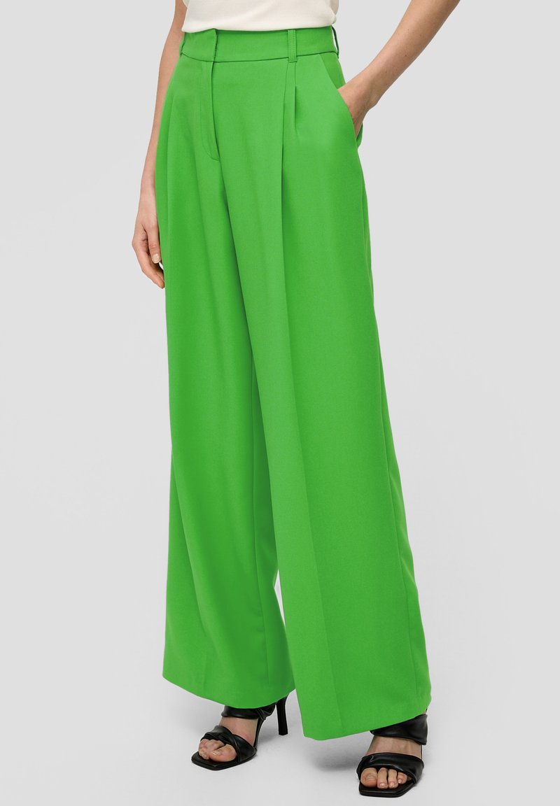 Pantalons larges verts vifs en tissu lisse, dotés de plis et de poches latérales. Portés avec des sandales à talons hauts noires à lanières.