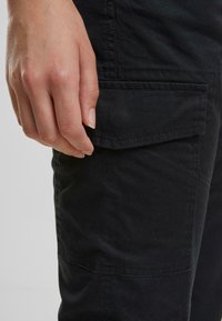 Svarte cargo-bukser har en sidelomme med klaff, dekorative sømmer og myk tekstur, holdt av en hånd nær stoffet.