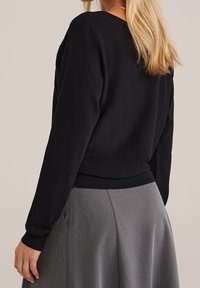 Pull noir avec poignets et ourlet côtelés, associé à une jupe grise. Le tissu semble doux avec une texture lisse. Des détails subtils sont visibles.