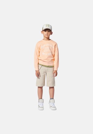 Felpa pesca con testo bianco, pantaloni corti khaki e sneakers bianche alte. Il ragazzo indossa un cappellino chiaro con dettagli verdi, in posizione eretta.