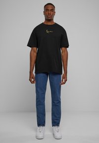 Black t-shirt with short sleeves and a gold logo on the chest, paired with blue jeans and white sneakers, set against a gray background.